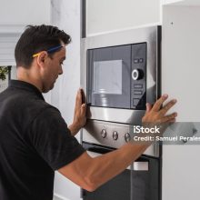 Caucasian adult male fitting a microwave inside the hole of a kitchen cabinet he is assembling for a customer.